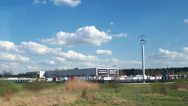 Ein Mercedes-Benz-Werk in Deutschland mit Fahrzeugen auf der Straße, umgeben von Gras, Pflanzen, Bäumen, Gebäuden und Pfählen, von denen einer ein Logo hat, unter einem bewölkten Himmel.