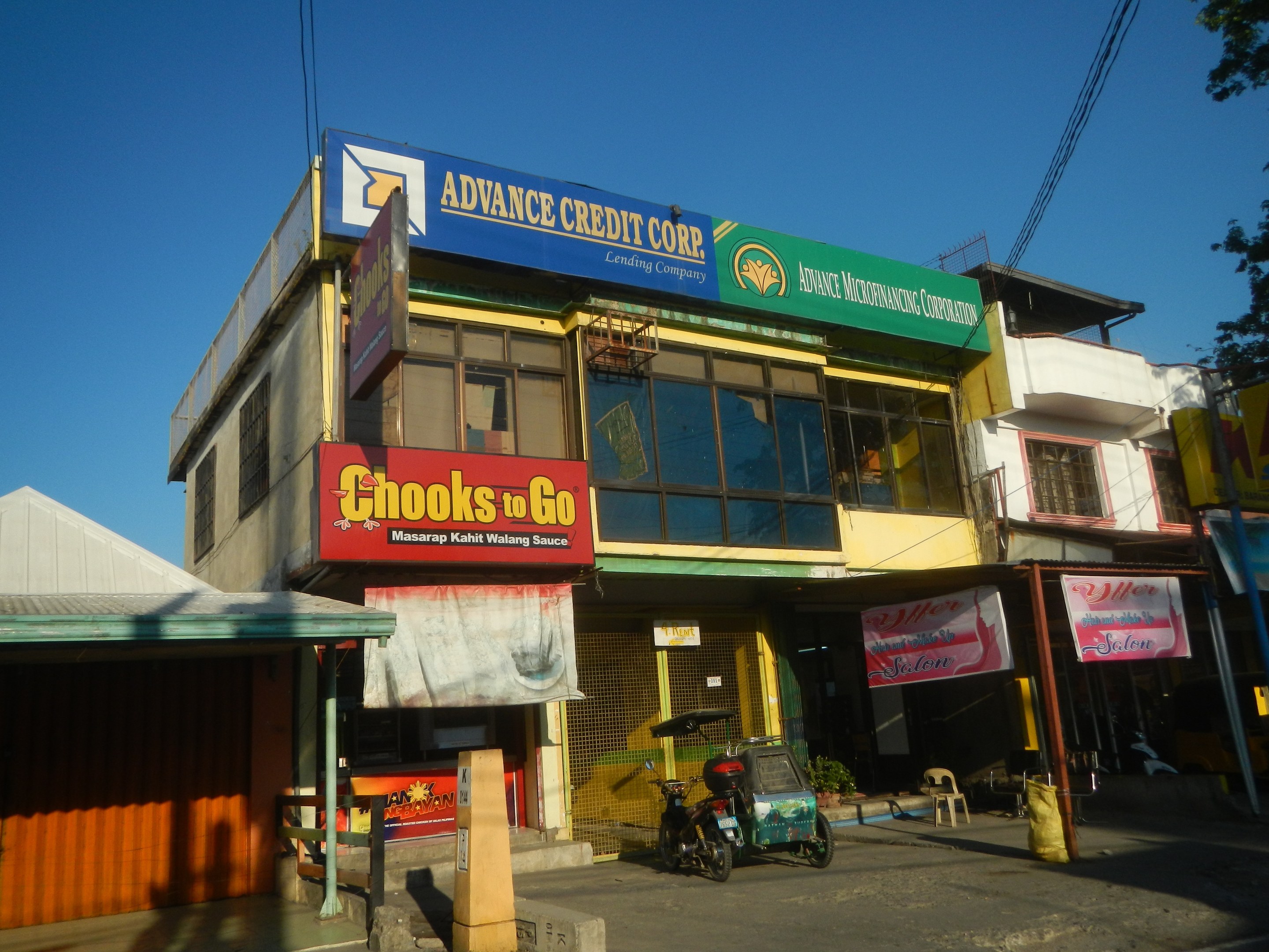 Exterior of a building with a sign reading "Advance Credit Corp" surrounded by vehicles, advertisement boards, electric poles, cables, trees, and a clear blue sky.