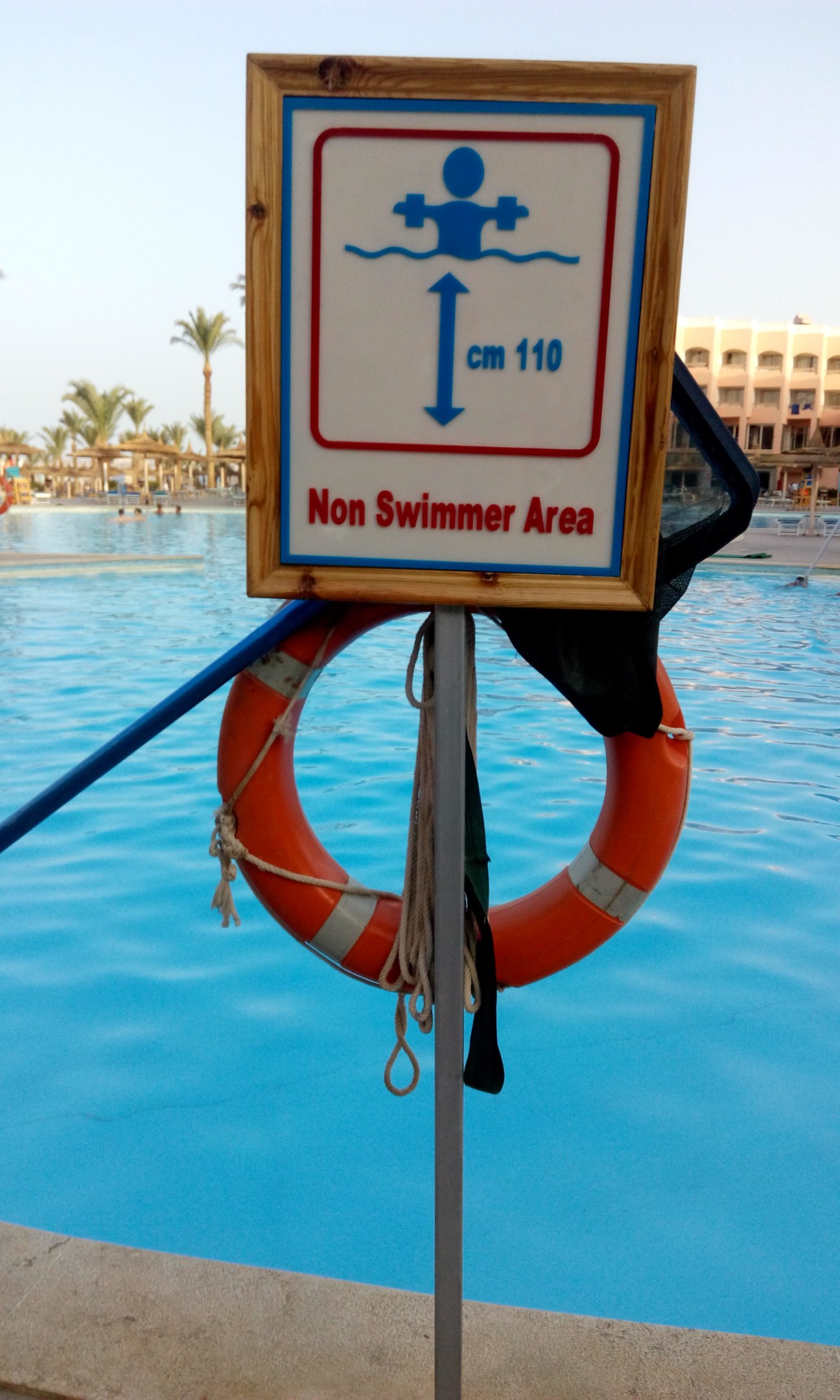 Schwimmbad mit einem "Nichtschwimmerbereich"-Schild und einem Rettungsring an einem Pfahl, umgeben von Bäumen, Gebäuden und einem klaren blauen Himmel.