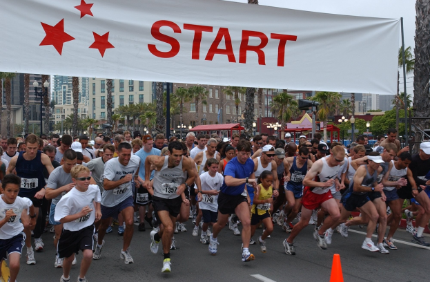 Gruppe von Menschen beim Marathonlauf mit einem Verkehrskegel im Vordergrund und einer Fahne im Hintergrund, umgeben von Bäumen, Laternenmasten, Gebäuden und einem klaren blauen Himmel.