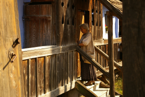 Eine ältere Frau steht auf den Stufen eines hölzernen Hauses mit einer hölzernen Tür auf der linken Seite und Gras auf dem Boden.