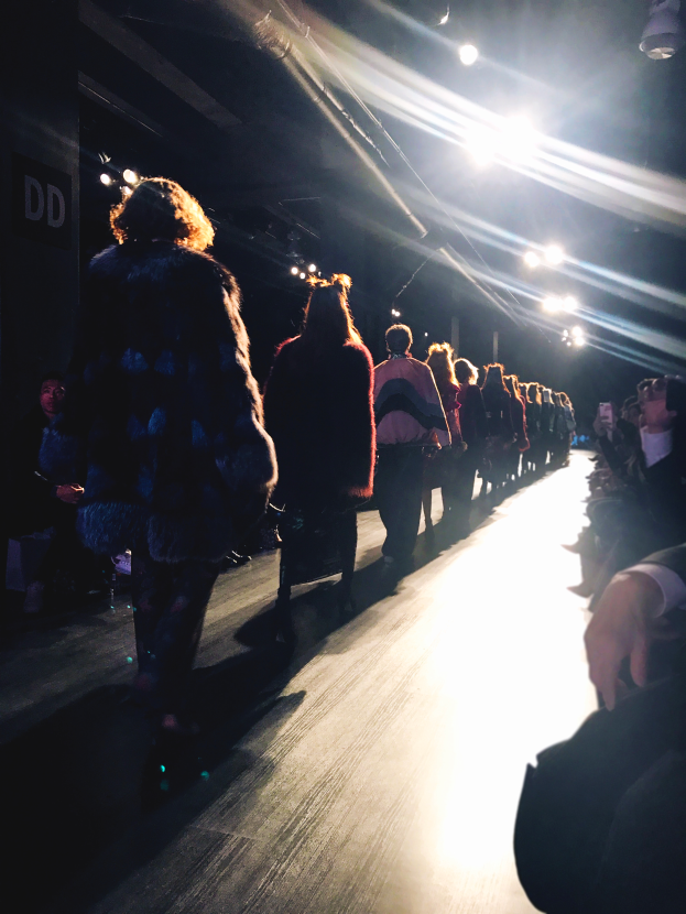 Eine Gruppe von Menschen läuft über den Laufsteg einer Modenschau, beleuchtet von hellen Scheinwerfern, mit einem Publikum auf beiden Seiten.