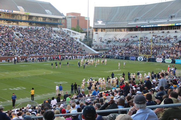Fußballspiel im Gange mit Spielern auf dem Feld und Zuschauern in den Rängen unter einem klaren blauen Himmel.