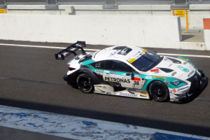 Ein Mercedes-AMG GT3 Rennwagen auf einer Rennstrecke, mit Text auf dem Auto und einer Wand mit Glasfenstern im Hintergrund.