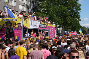 Eine große Menschenmenge marschiert auf der Straße bei der Berlin Pride 2019, viele tragen Mützen und Schutzbrillen, halten Fahnen und Schilder, mit Gebäuden im Hintergrund unter einem bewölkten Himmel.