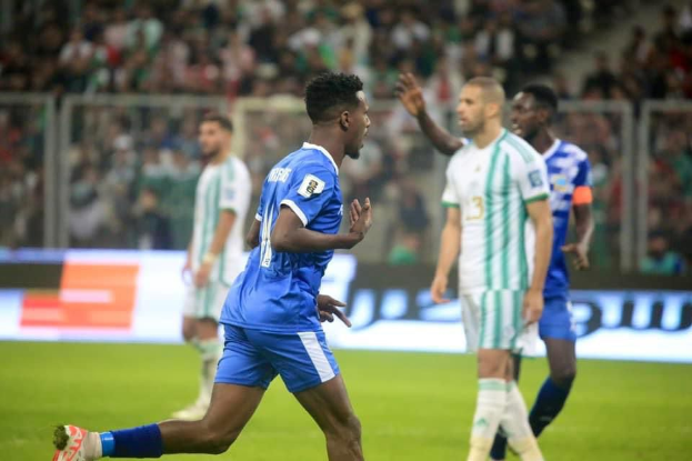 Ein Fußballspieler in einem blauen und weißen Trikot feiert nach einem Tor, umgeben von einer Gruppe von Menschen in weißen und grünen Trikots, mit einer weißen und blauen Tafel und einem Geländer im Hintergrund.