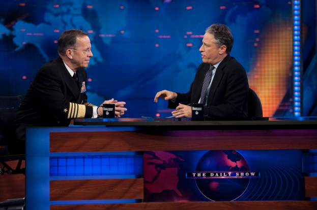 Zwei Manner in schwarzen Blazern, Hemden und Krawatten sitzen an einem Tisch mit Mikrofonen zwischen ihnen vor einem Fernseher und einem blauen Hintergrund.