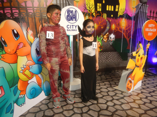 Zwei Kinder in Halloween-Kostümen stehen nebeneinander vor einer Wand mit einem Gemälde eines Gebäudes, Bäume, Laternenmasten, einem Zaun und Schildern mit Text und Tierbildern.