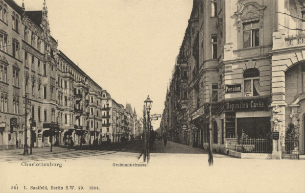 Schwarzes und weißes Foto einer Stadtstraße in Charlottenburg, Deutschland, mit Gebäuden, Straßenlaternen, Schildern, Motorfahrzeugen, Fußgängern, Gittern und einem klaren Himmel.