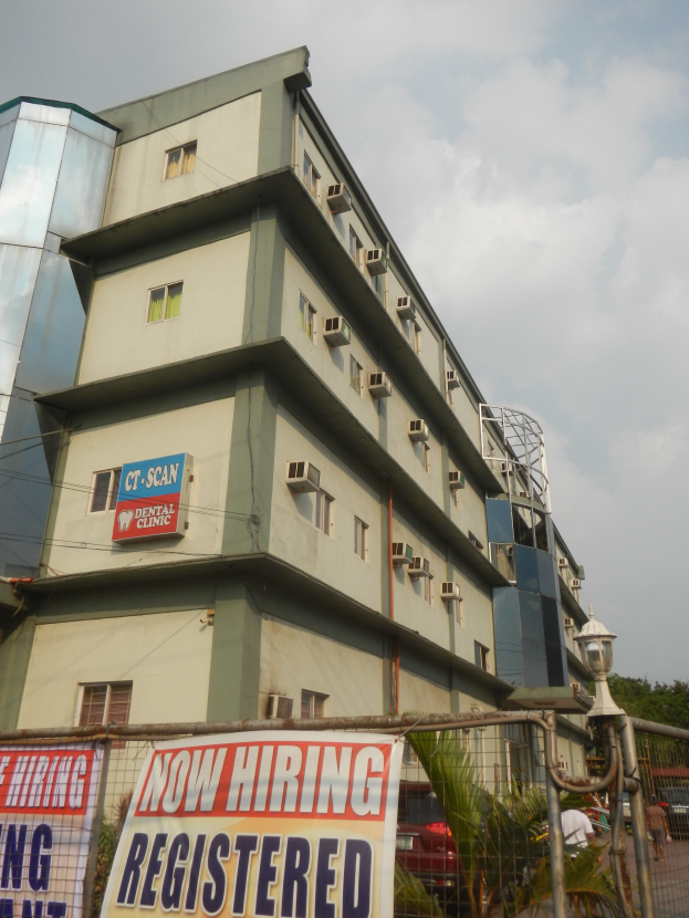 Ein Gebäude mit Fenstern, Klimaanlagen und einem "Jetzt Einstellen Registriert"-Schild, umgeben von einem Metallzaun, einer Straßenlaterne, Pflanzen, Bäumen, Fahrzeugen, unter einem bewölkten Himmel.