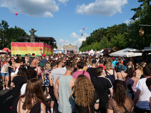 Eine große Menschenmenge, die eine Straße mit Zelten, Bäumen, Pfählen, Lichtern und einer Statue gesäumt, im Hintergrund Gebäude und ein Himmel mit Wolken und Ballons, während des Christopher Street Day in Berlin.