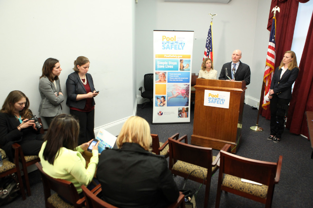 Drei Frauen sitzen auf Stühlen, während ein Mann und eine Frau an einem Rednerpult in der Mitte stehen, mit zwei Fahnen und einem Banner im Hintergrund; links sieht eine Frau auf eine digitale Kamera, während zwei andere Frauen stehen und Vorhänge in der oberen rechten Ecke zu sehen sind.