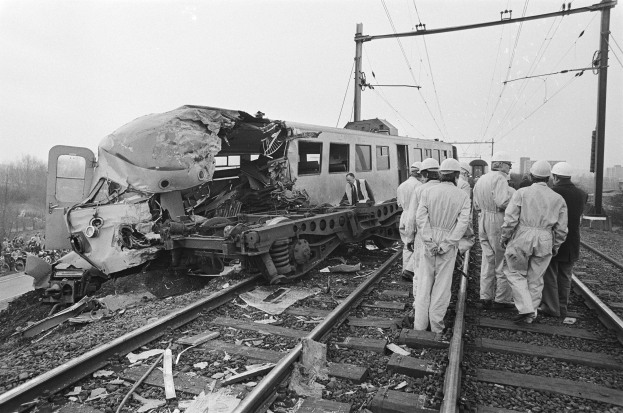 Schwarz-weiß-Bild einer Zugkollision auf den Schienen mit Einsatzkräften in Helmen und einem Hintergrund aus Strommasten, Bäumen, Gebäuden und einem klaren Himmel.
