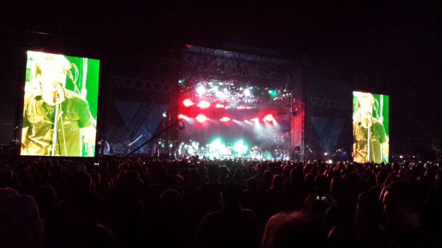 Ein Konzertszene mit einer Menge unten, ein großer Bildschirm links und ein kleiner Bildschirm rechts.