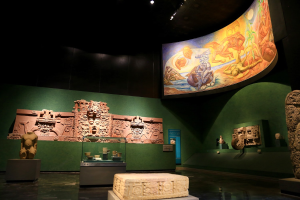 Ausstellungsraum im Anthropologischen Museum von Mexiko-Stadt mit verschiedenen Artefakten wie Skulpturen, Tischobjekten, einer Texttafel, einer Wandmalerei und Deckenbeleuchtung.