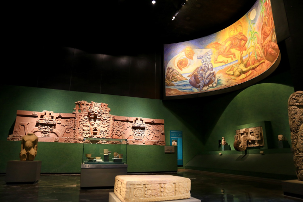 Ausstellungsraum im Anthropologischen Museum von Mexiko-Stadt mit verschiedenen Artefakten wie Skulpturen, Tischobjekten, einer Texttafel, einer Wandmalerei und Deckenbeleuchtung.