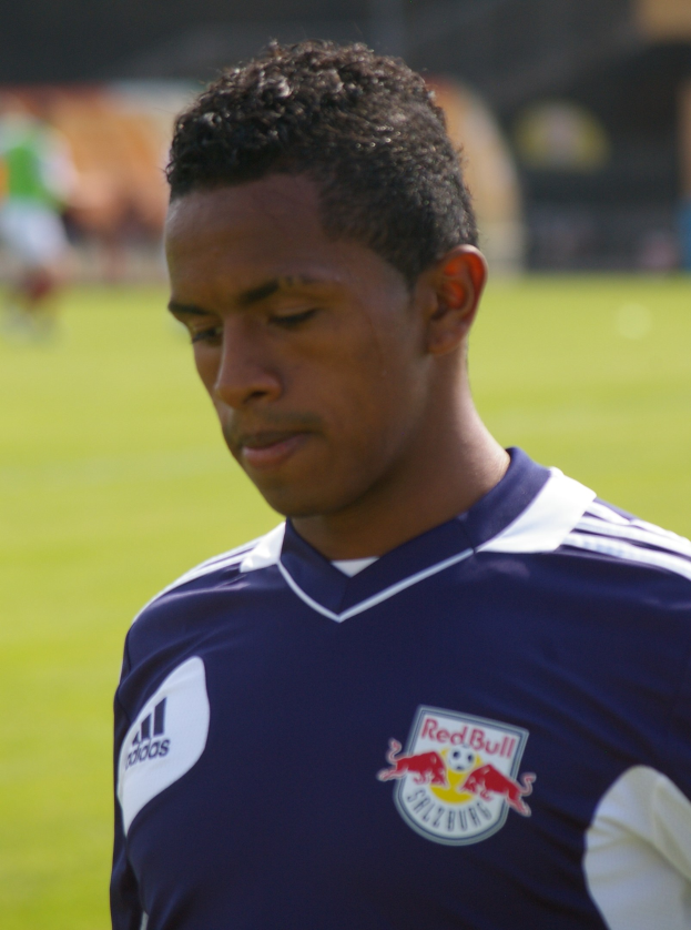 Ein junger Mann in einem blauen T-Shirt steht auf einem Fußballfeld mit einem unscharfen Hintergrund, er wirkt wie ein Flügelstürmer mit kurzen dunklen Haaren und einem entschlossenen Gesichtsausdruck.