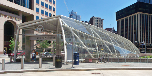Eine belebte Stadtstraße mit hohen Wolkenkratzern, ein Bushaltestelle mit Glasdach, Menschen, Fahrzeuge, ein Metallzaun, ein Müllcontainer, Pfosten, ein Schild, eine Verkehrsampel, Bäume, ein Kran und ein bewölkter Himmel.