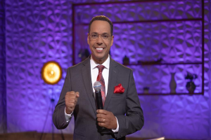 Stephen Colbert, der eine Anzug, Krawatte und eine Brille trägt, steht auf einer Bühne und hält ein Mikrofon in der Hand, mit einer Wand und verschiedenen Gegenständen dahinter.