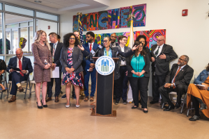 Eine Gruppe von Menschen steht vor einem Podium mit einem Mikrofon und einer Tafel mit der Aufschrift "Sancho Stadtrat tritt dem Stadtrat bei", mit anderen im Hintergrund sitzend und stehend, Flaggen, eine Tafel, einen Feuerlöscher und Deckenleuchten an der Wand.