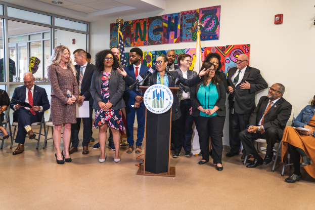 Eine Gruppe von Menschen steht vor einem Podium mit einem Mikrofon und einer Tafel mit der Aufschrift "Sancho Stadtrat tritt dem Stadtrat bei", mit anderen im Hintergrund sitzend und stehend, Flaggen, eine Tafel, einen Feuerlöscher und Deckenleuchten an der Wand.