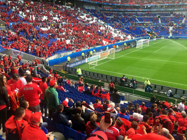 Eine große Menge Menschen in einem Stadion schaut einem Fußballspiel zu, mit Sitzplätzen und Stehplätzen, Bannern, einem Torpfosten und Deckenleuchten.