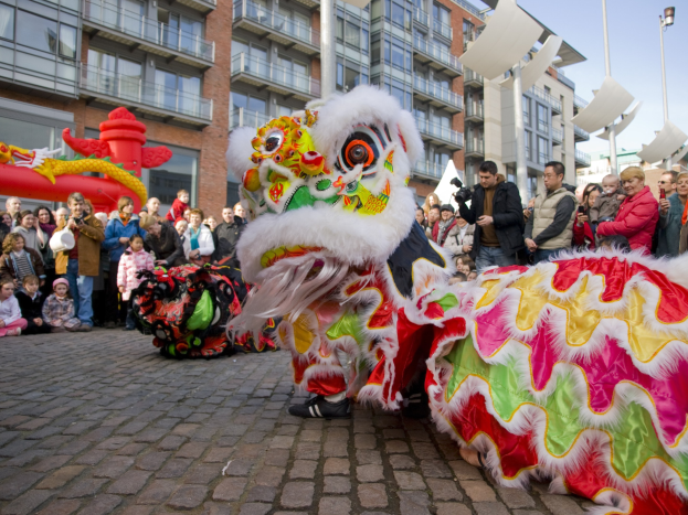 Ein lebendiges chinesisches Neujahrsfest in Amsterdam mit einer Drachenvorführung vor einer Zuschauermenge, einige halten Kameras, vor einem Hintergrund aus Gebäuden, Laternenmästen und einem klaren blauen Himmel.