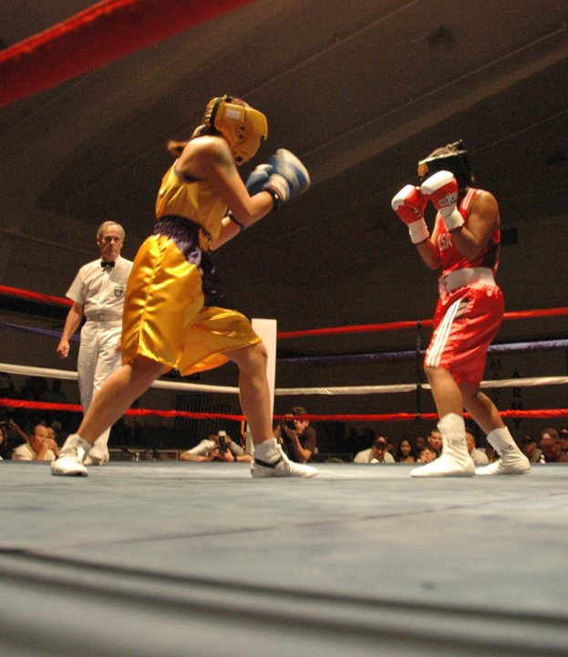 Zwei Boxer im Ring, die sich gegenseitig boxen, während ein Schiedsrichter im Hintergrund zusieht, umgeben von Fotografen und Zuschauern unter einer sichtbaren Decke.
