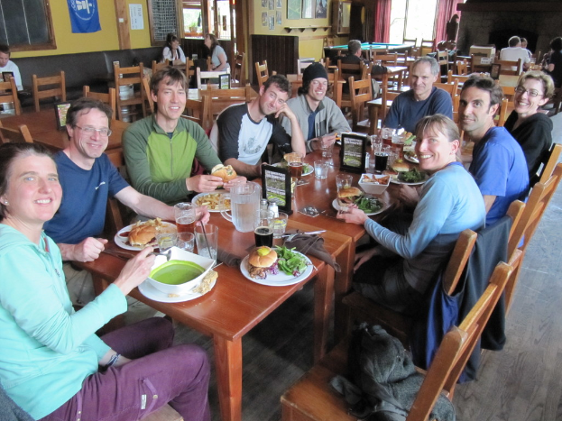 Eine Gruppe von Menschen sitzt um einen Tisch mit Tellern, Schalen, Gl√§sern, Flaschen, einem Wasserglas, Karten, Tuch und Essen, lachend, mit einer Jacke auf einem der Stühle, w√§hrend einige weitere Personen an anderen Tischen sitzen, mit einem Banner, Bildern und Postern an der Wand und einem Fenster mit Vorh√§ngen.