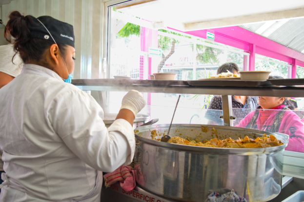 Eine Frau in Kochuniform, weißem Kleid, Handschuhen und schwarzer Mütze, hält einen Löffel und steht vor einer Stahlschüssel mit Essen, mit einer anderen Frau in einem blauen Kleid links von ihr und verschiedenen Küchenutensilien und Menschen im Hintergrund.
