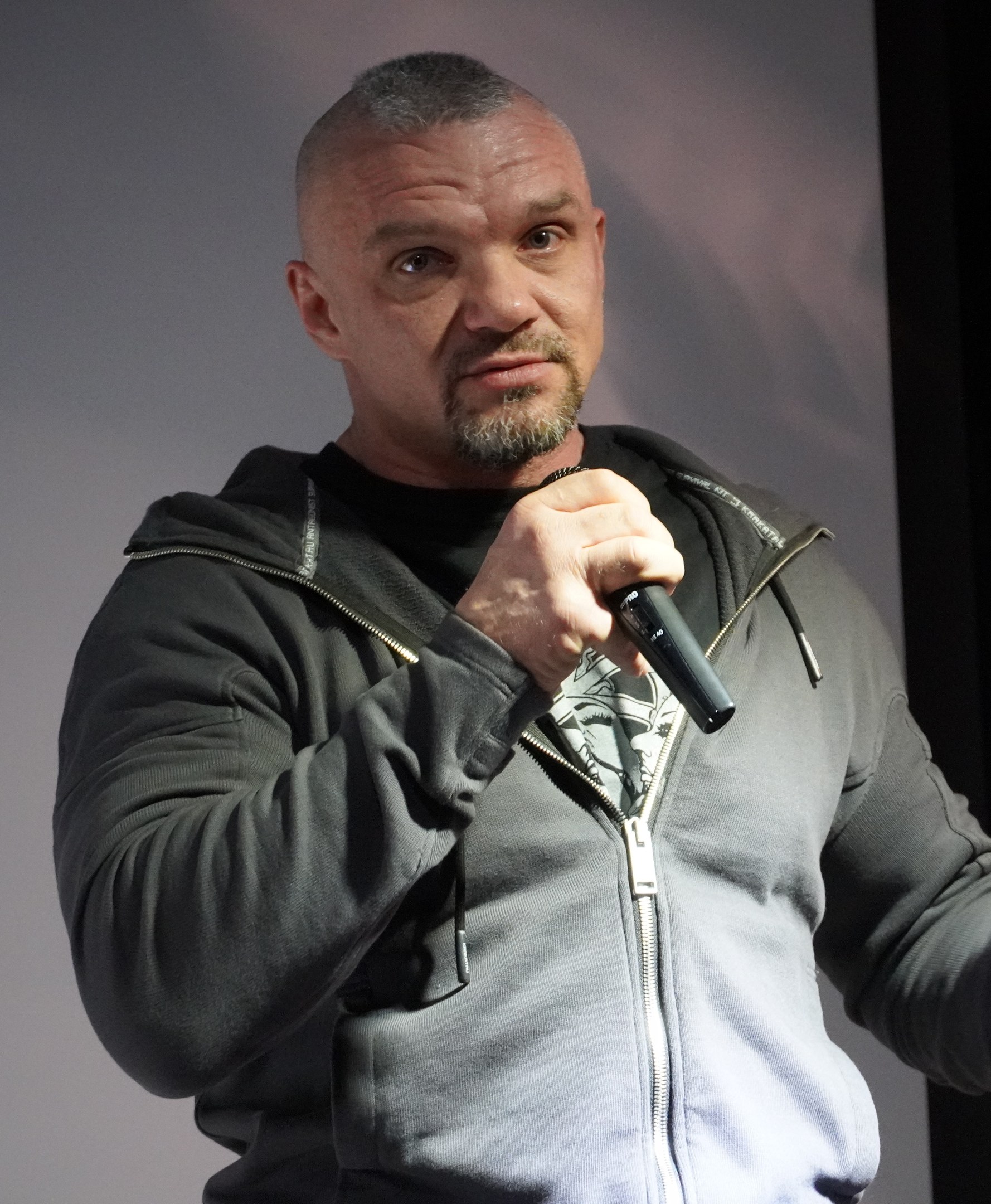 Ein Mann in einer schwarzen Kapuzenjacke und einem schwarzen Anzug, der ein weißes Hemd trägt und mit zurückgekämmtem Haar selbstbewusst vor einer Wand steht und ein Mikrofon hält.