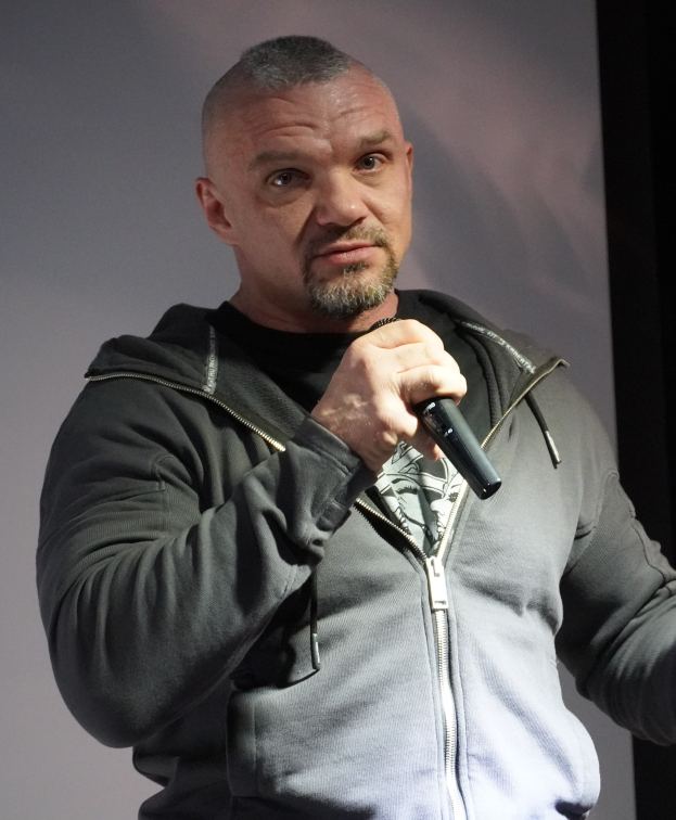 Ein Mann in einer schwarzen Kapuzenjacke und einem schwarzen Anzug, der ein weißes Hemd trägt und mit zurückgekämmtem Haar selbstbewusst vor einer Wand steht und ein Mikrofon hält.