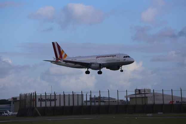 Germanwings Airbus A320-200 beim Start von Frankfurt Airport, mit Gras darunter, einem Zaun im Vordergrund, Gebäuden und Bäumen im Hintergrund und einer bewölkten Himmel