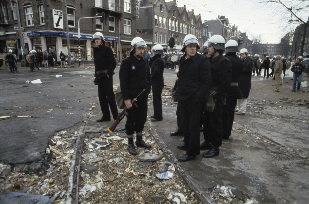 Eine Gruppe von Polizisten in Helmen und mit Waffen in der Hand steht an der Seite einer Straße. Im Hintergrund sind Gebäude, Schilder, Laternenpfähle, Bäume und ein klarer Himmel zu sehen.