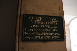 Eine Tafel an einer Gebäudewand trägt die Aufschrift "Chapel Royal Sunday Services Visitors Welcome", mit einer Säule rechts daneben.