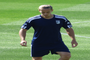 Ein Mann in einem blauen T-Shirt und Shorts schreitet selbstbewusst über ein grünes Feld und sieht aus wie ein professioneller Fussballspieler mit entschlossener Miene.
