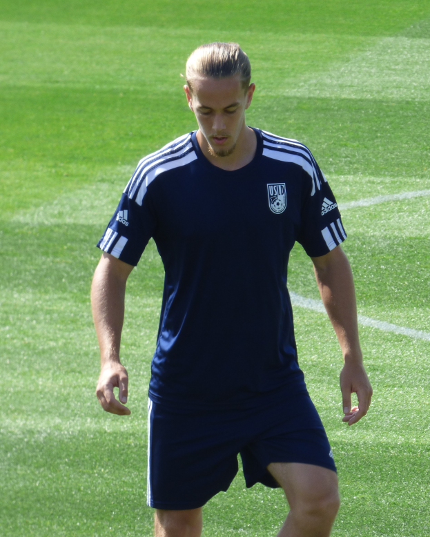 Ein Mann in einem blauen T-Shirt und Shorts schreitet selbstbewusst über ein grünes Feld und sieht aus wie ein professioneller Fussballspieler mit entschlossener Miene.