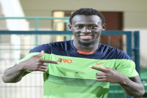 Ein Mann in einem grün-blauen Hemd, identifiziert als Emmanuel Adebayor, zeigt mit einem Lächeln auf die Kamera, mit einem unscharfen Geländer, Wand und Tür im Hintergrund.