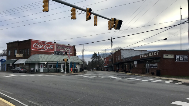 Stadtstraßenkreuzung mit Ampel, Fahrzeugen, Gebäuden mit Beschilderung, Strommasten und -leitungen, Bäumen und einem bewölkten Himmel.