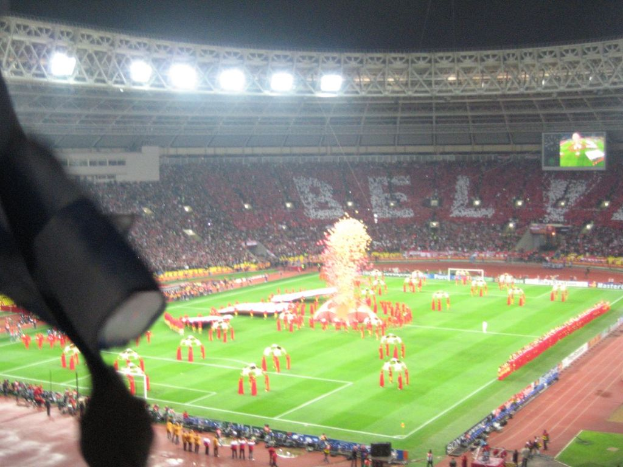 Ein Stadion mit Flutlicht, Eisengittern, Zuschauern, stehenden Individuen, Werbetafeln, Anzeigetafeln, einer Kamera und Fahnen und Dekorationen.