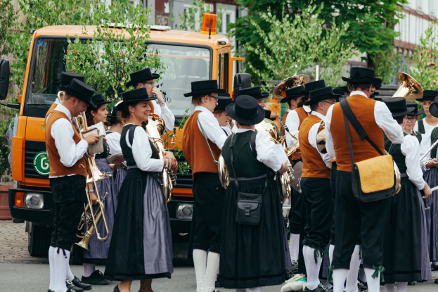 Eine Gruppe von Menschen in traditioneller bayrischer Tracht, die eine Straße entlangmarschieren, Instrumente spielen und Taschen tragen, mit Bäumen, Gebäuden und einem geparkten Fahrzeug im Hintergrund.