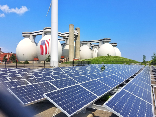Solarpanels auf dem Dach eines Gebäudes mit einer Flagge, umgeben von einem Zaun, Gras, Pflanzen, Bäumen und anderen Objekten, sowie zusätzlichen Gebäuden und einem bewölkten Himmel im Hintergrund.