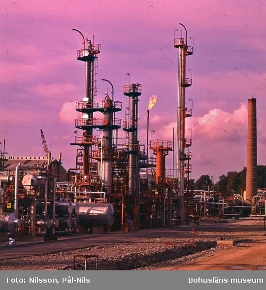 Ölraffinerie mit ausgedehnten Rohrleitungen, Metallstrukturen, Containern, Bäumen, einem Gebäude, einem Kran und einem bewölkten Himmel, einschließlich Text unten.