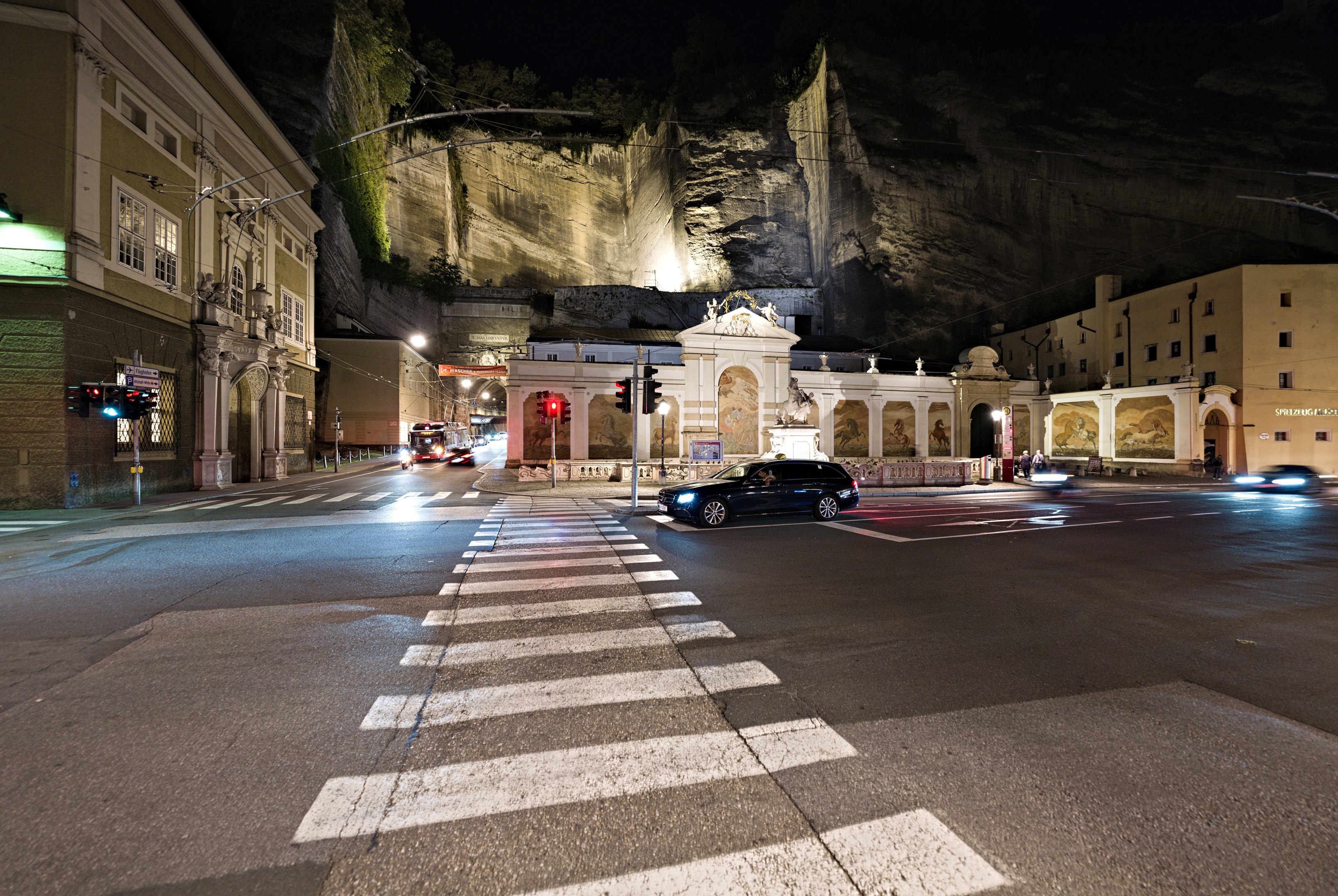Eine nächtliche Stadtstraße mit fahrenden Autos, Straßenlaternen, Ampeln, beleuchteten Gebäuden, Laternenmasten, einem Hügel im Hintergrund und einer Zebrastreifen am Boden.