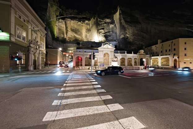 Eine nächtliche Stadtstraße mit fahrenden Autos, Straßenlaternen, Ampeln, beleuchteten Gebäuden, Laternenmasten, einem Hügel im Hintergrund und einer Zebrastreifen am Boden.