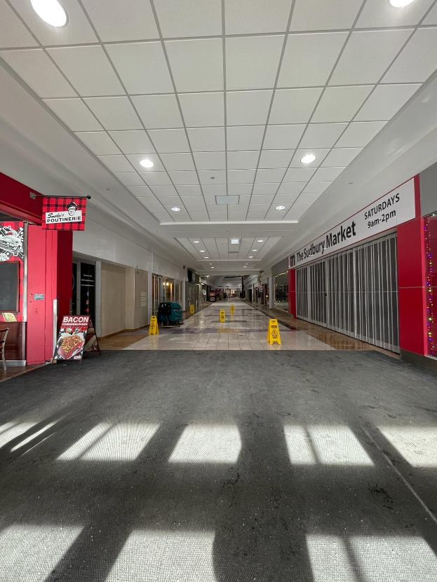 Innenansicht eines hell erleuchteten Einkaufszentrums mit einem Stuhl und einer Tafel, auf der "Sydbury Market - Vorher und Nachher" steht, links, und verstreuten Gegenständen rechts und auf dem Boden.