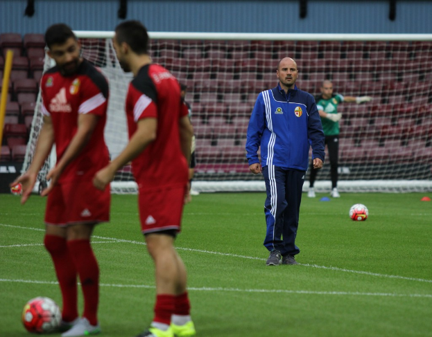 Ein Fußballfeld mit darauf stehenden Spielern, zwei Fußball auf dem Boden und einer leeren Tribüne im Hintergrund.