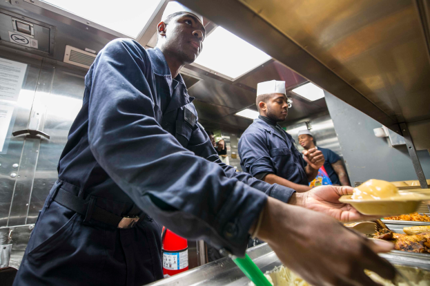 Eine Gruppe Von M├Ąnnern Steht In Einer K├╝che, Einer Hält Ein Tablett Mit Speisen, Mit Tabletts Von Speisen Auf Dem Tisch, Einem Feuerl├Âscher Und Papieren An Der Wand Im Hintergrund.