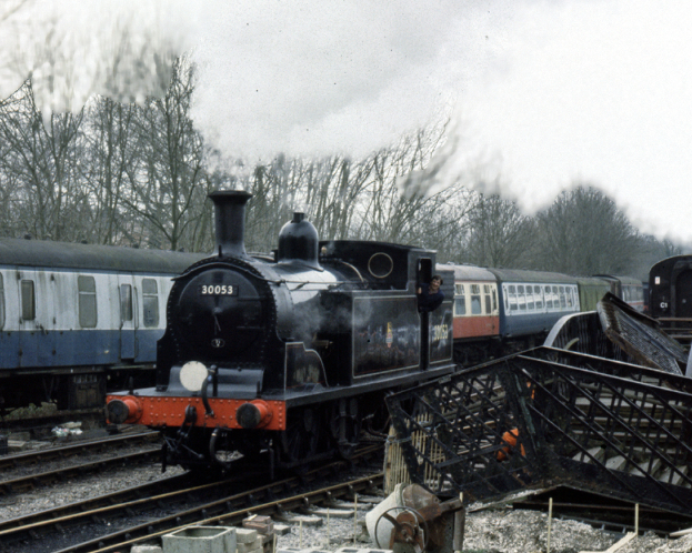 Ein Motor bewegt sich auf einer Bahn mit einem Zug dahinter, während einige Objekte seitlich im Vordergrund platziert sind, mit Bäumen und Himmel im Hintergrund.