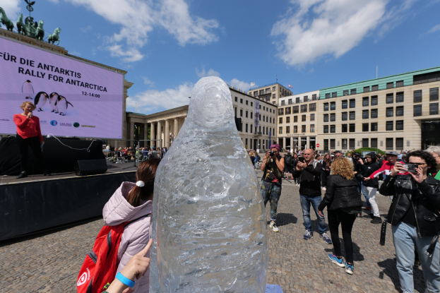 Eine Gruppe von Menschen versammelt sich um eine große Eisskulptur vor einem Gebäude, während eine Person auf der linken Seite der Bühne mit einem Mikrofon spricht und von Lautsprechern und einer Wand umgeben ist, während andere die Szene fotografieren, die von Gebäuden und einem bewölkten Himmel umgeben ist.